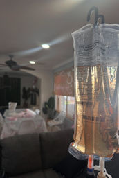 IV saline drip bag (0.9% sodium chloride) hanging in a home setting with a cozy living room background — sofa, dining table, window blinds and ceiling fan softly blurred.