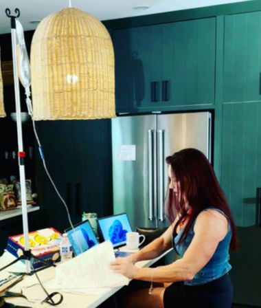 Person working at a kitchen island with papers and two laptops, coffee mug on the counter, woven rattan pendant light, dark green cabinets and stainless-steel refrigerator, IV pole visible nearby