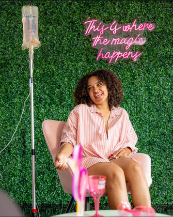 Smiling woman in a pink striped shirt relaxes in a pink chair receiving an IV infusion, green hedge wall backdrop and pink neon sign reading 'This is where the magic happens' in a wellness lounge