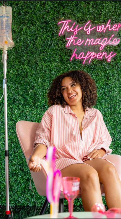Smiling woman in a pink striped shirt relaxes in a pink chair receiving an IV infusion, green hedge wall backdrop and pink neon sign reading 'This is where the magic happens' in a wellness lounge