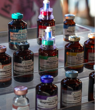 Amber glass IV vitamin vials with colorful caps (labels show Vitamin D3, Glutathione, L‑Carnitine) neatly arranged on a clear display shelf at a wellness clinic — intravenous nutrient therapy.