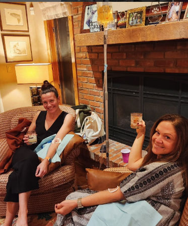 Two women relaxing in a cozy living room by a brick fireplace, receiving IV hydration therapy and toasting with drinks