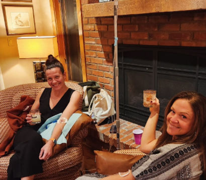 Two women relaxing in a cozy living room by a brick fireplace, receiving IV hydration therapy and toasting with drinks
