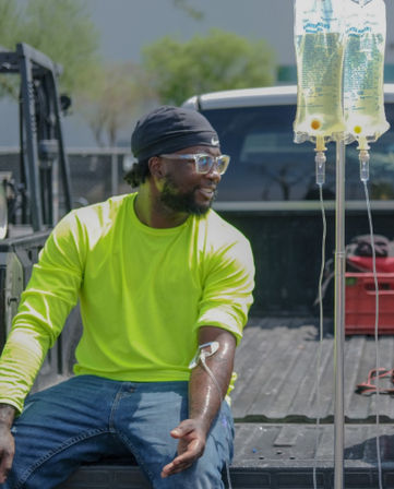Worker in a neon safety shirt and beanie sits on a pickup truck bed at an outdoor worksite, receiving IV fluids from a mobile IV pole.