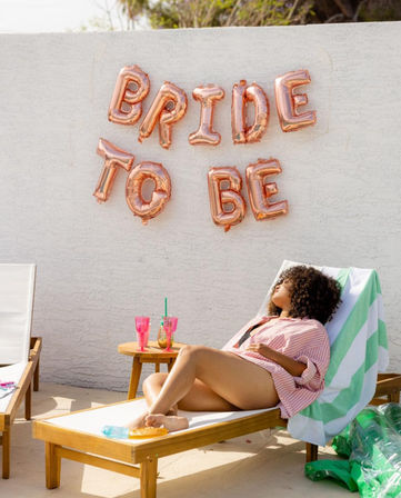 Rose-gold foil balloons spelling "BRIDE TO BE" on a white stucco wall above a person lounging on a poolside sun lounger with a green-striped towel, pink drinks on a wooden side table and summer accessories.