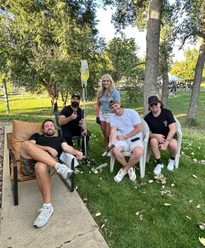 Five adults relaxing on chairs in a sunny park under trees at an outdoor event, smiling around an IV hydration stand with tents and people visible in the background.