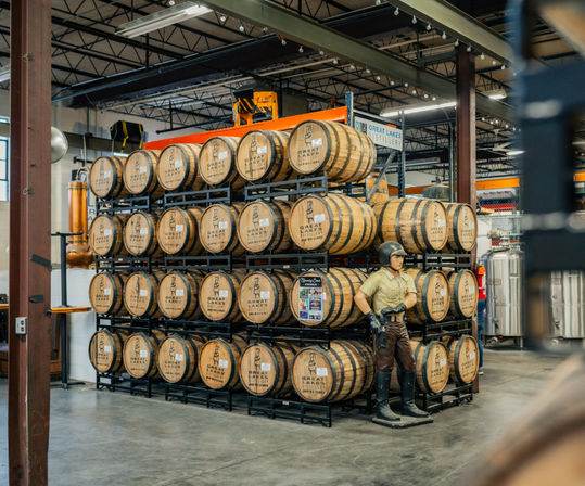 Stacked oak barrels on metal racks inside a bright industrial distillery warehouse, with a vintage aviator mannequin posed beside the barrels and stainless steel tanks in the background.