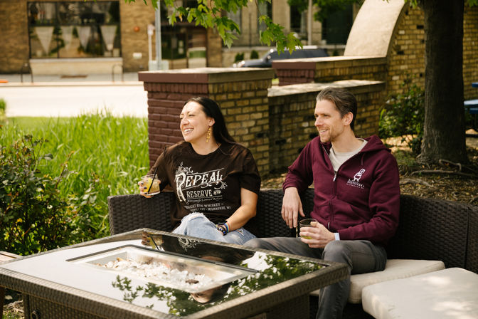 Two friends laughing with drinks on a shaded downtown outdoor patio lounge with wicker seating, glass-topped fire-pit table, and brick wall backdrop