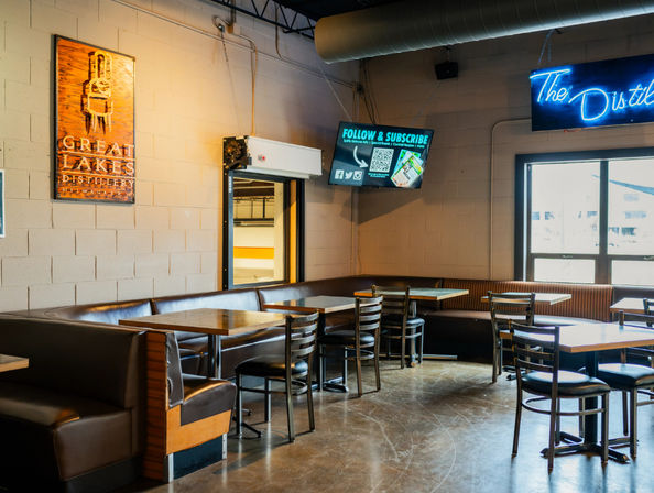 Industrial-style distillery tasting room with brown leather booths, wooden tables and metal chairs on a polished concrete floor, exposed ductwork, a neon wall sign, wall-mounted TV and large windows letting in daylight.