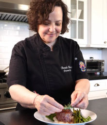 Smiling chef in a black jacket carefully garnishing a plated steak with rosemary and asparagus in a bright modern white kitchen.