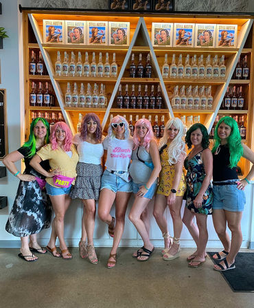 Eight women in colorful wigs and summer outfits posing together inside an Austin tasting room in front of triangular wooden shelves stocked with liquor bottles — playful bachelorette-style group photo.