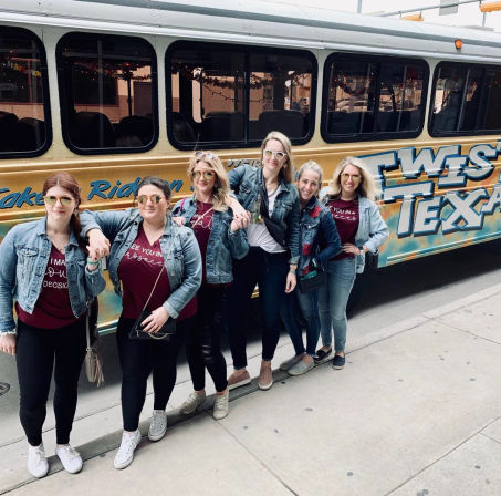 Six women in denim jackets and maroon shirts smiling and posing on a city sidewalk in front of a yellow shuttle bus with large "West Texas" lettering