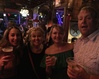 Four friends smiling and holding drinks in a dim, rustic bar with neon lights, wooden walls and a lively nighttime nightlife vibe.