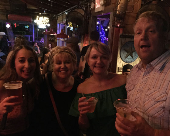 Four friends smiling and holding drinks in a dim, rustic bar with neon lights, wooden walls and a lively nighttime nightlife vibe.