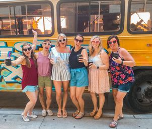 Six friends in summer outfits and heart-shaped sunglasses holding drinks and posing in front of a yellow decorated party bus on a sunny city sidewalk, celebrating a bachelorette.