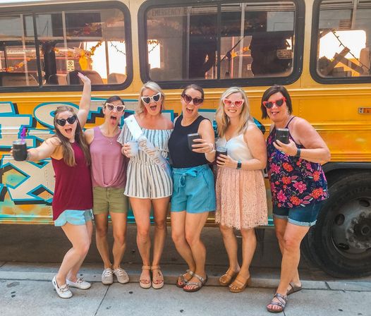 Six friends in summer outfits and heart-shaped sunglasses holding drinks and posing in front of a yellow decorated party bus on a sunny city sidewalk, celebrating a bachelorette.