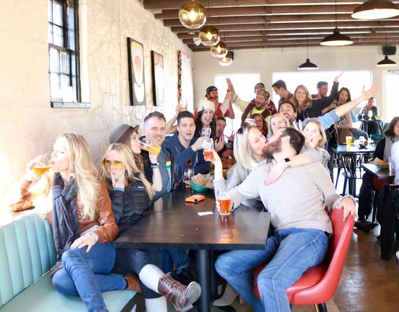 Sunlit taproom scene with a lively group of friends at a long table enjoying craft beers, cheering, laughing and sharing a kiss under brass pendant lights.