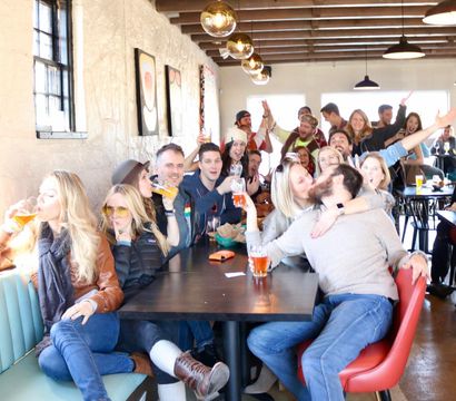 Sunlit taproom scene with a lively group of friends at a long table enjoying craft beers, cheering, laughing and sharing a kiss under brass pendant lights.