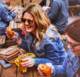 Two friends in denim jackets laughing at an outdoor beer garden patio, holding craft beers and pretzel sandwiches at a wooden picnic table