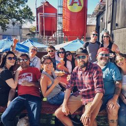 Group of friends at a craft brewery beer garden patio, seated at a red picnic table under blue umbrellas, enjoying beer flights with large red fermentation tanks in the background on a sunny day.