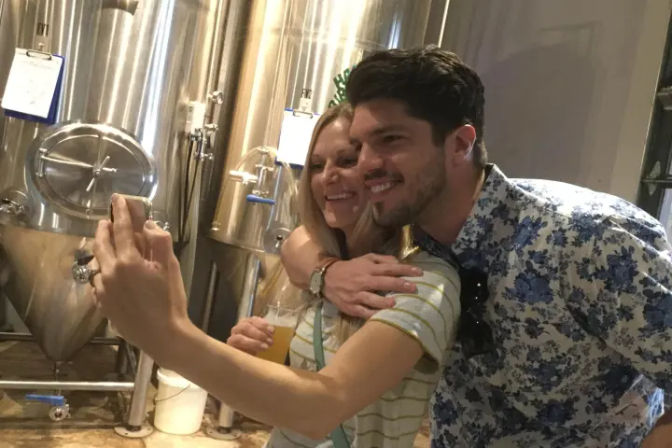 Two smiling people taking a selfie with beers in hand inside a craft brewery, posing in front of shiny stainless-steel fermenters