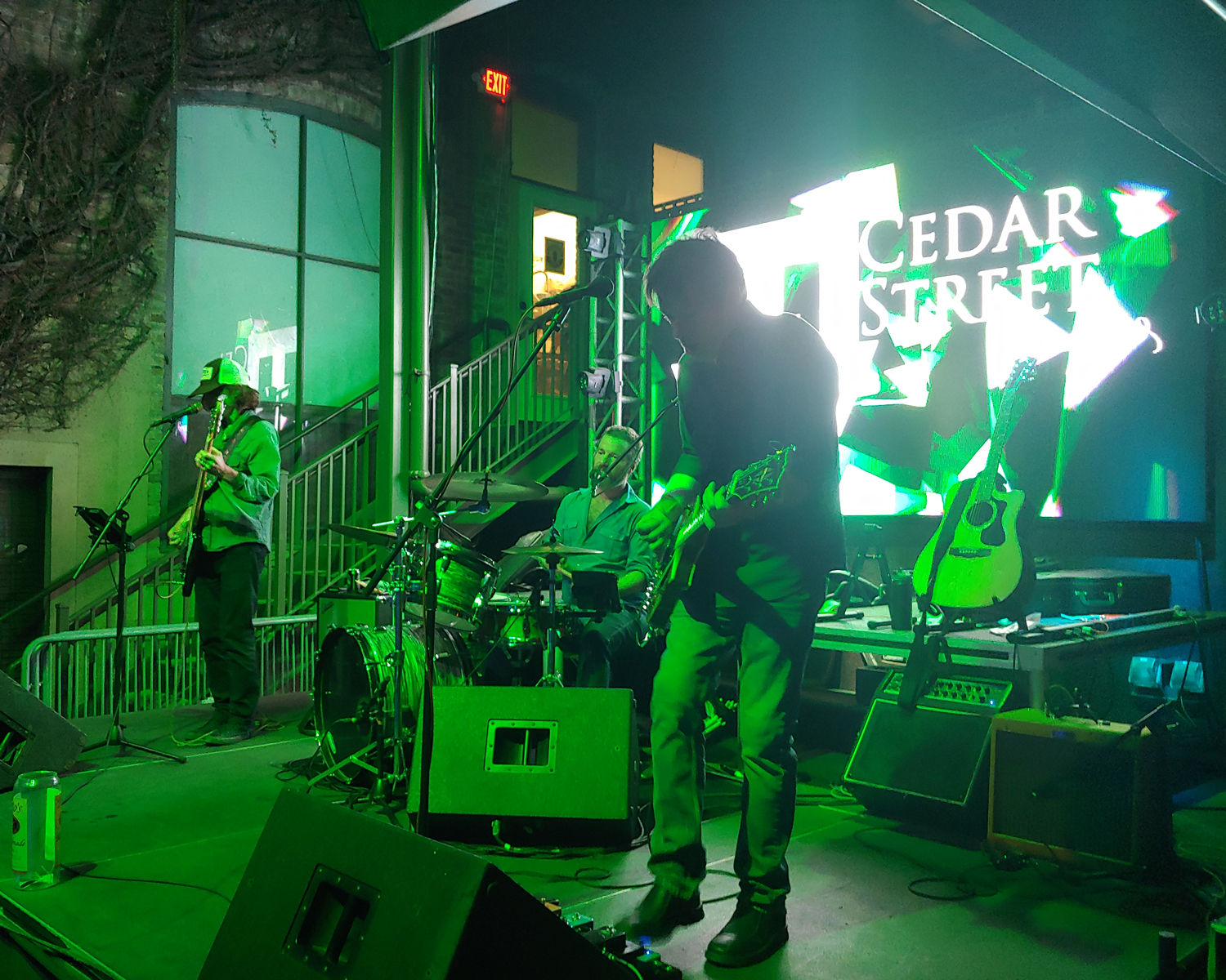 Three-piece live band performing on an outdoor night stage bathed in green light — guitarist mid-riff, drummer, bassist, LED backdrop and gear