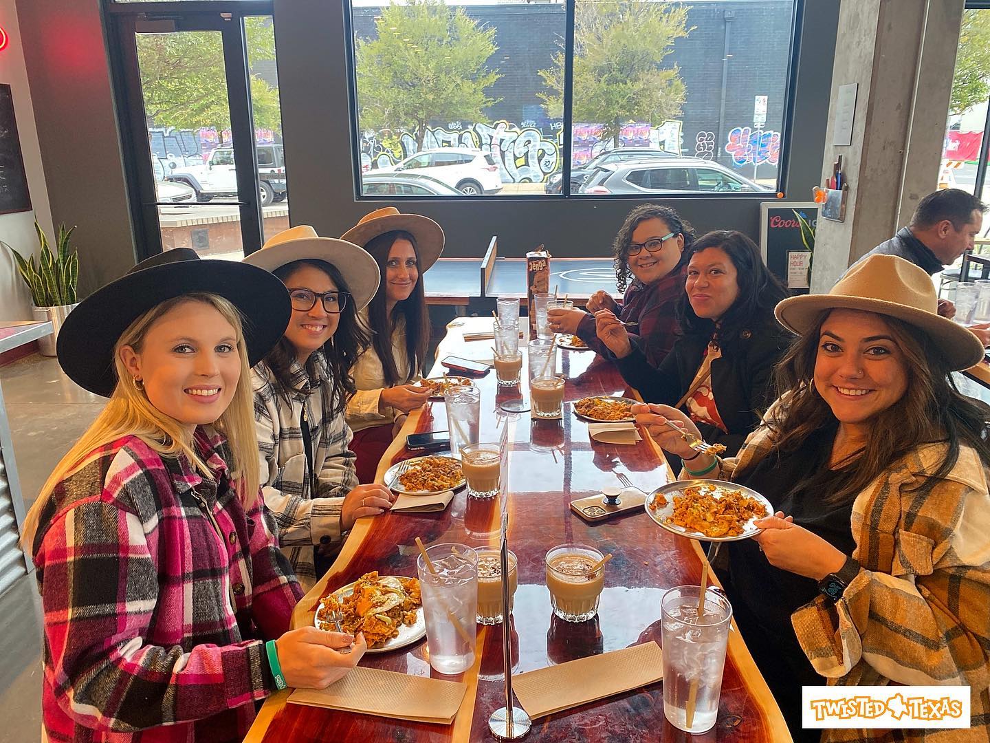 Smiling group of friends in hats and flannel sharing brunch at a long wooden table with plates of loaded nachos, iced coffees, and large windows showing street art in an urban cafe.