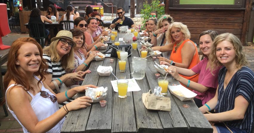 Large group of friends enjoying brunch outdoors at a rustic wooden picnic table, smiling and sharing foil-wrapped tacos and glasses of orange juice on a sunny patio