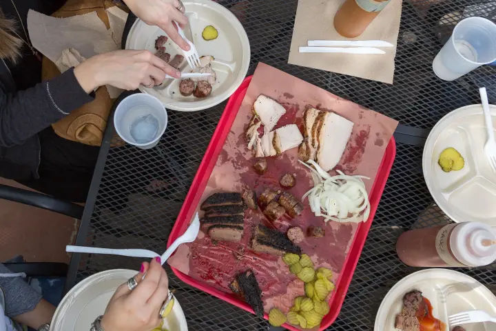 Overhead shot of an outdoor metal table with a red tray of smoked BBQ — sliced brisket, sausage links, smoked turkey, pickles and sliced onion — surrounded by paper plates, plastic utensils, sauce bottle and diners’ hands, al fresco barbecue meal.