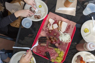 Overhead shot of an outdoor metal table with a red tray of smoked BBQ — sliced brisket, sausage links, smoked turkey, pickles and sliced onion — surrounded by paper plates, plastic utensils, sauce bottle and diners’ hands, al fresco barbecue meal.