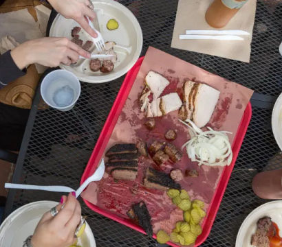 Overhead shot of an outdoor metal table with a red tray of smoked BBQ — sliced brisket, sausage links, smoked turkey, pickles and sliced onion — surrounded by paper plates, plastic utensils, sauce bottle and diners’ hands, al fresco barbecue meal.