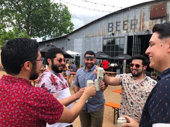 Group of friends toasting canned beers at an outdoor beer garden with a rustic corrugated-metal building, string lights, umbrellas and picnic tables