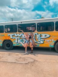 Two women jumping and posing in front of a colorful painted Texas tour bus with graffiti-style lettering under a bright summer sky