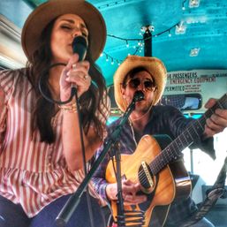 Acoustic duo performing inside a decorated bus — woman in a wide-brim hat and striped blouse singing into a mic, man in a straw hat and sunglasses playing an acoustic guitar, string lights and turquoise ceiling overhead.