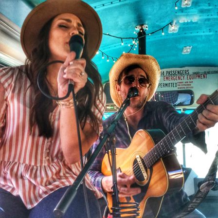 Acoustic duo performing inside a decorated bus — woman in a wide-brim hat and striped blouse singing into a mic, man in a straw hat and sunglasses playing an acoustic guitar, string lights and turquoise ceiling overhead.