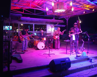 Four-piece live band on an outdoor wooden stage at night under purple-pink lights — electric guitar, bass, drum kit, and saxophonist with a "Tip the Band" sign.