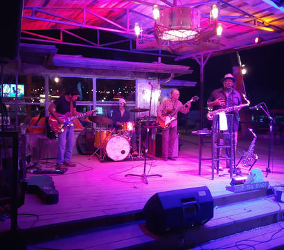 Four-piece live band on an outdoor wooden stage at night under purple-pink lights — electric guitar, bass, drum kit, and saxophonist with a "Tip the Band" sign.
