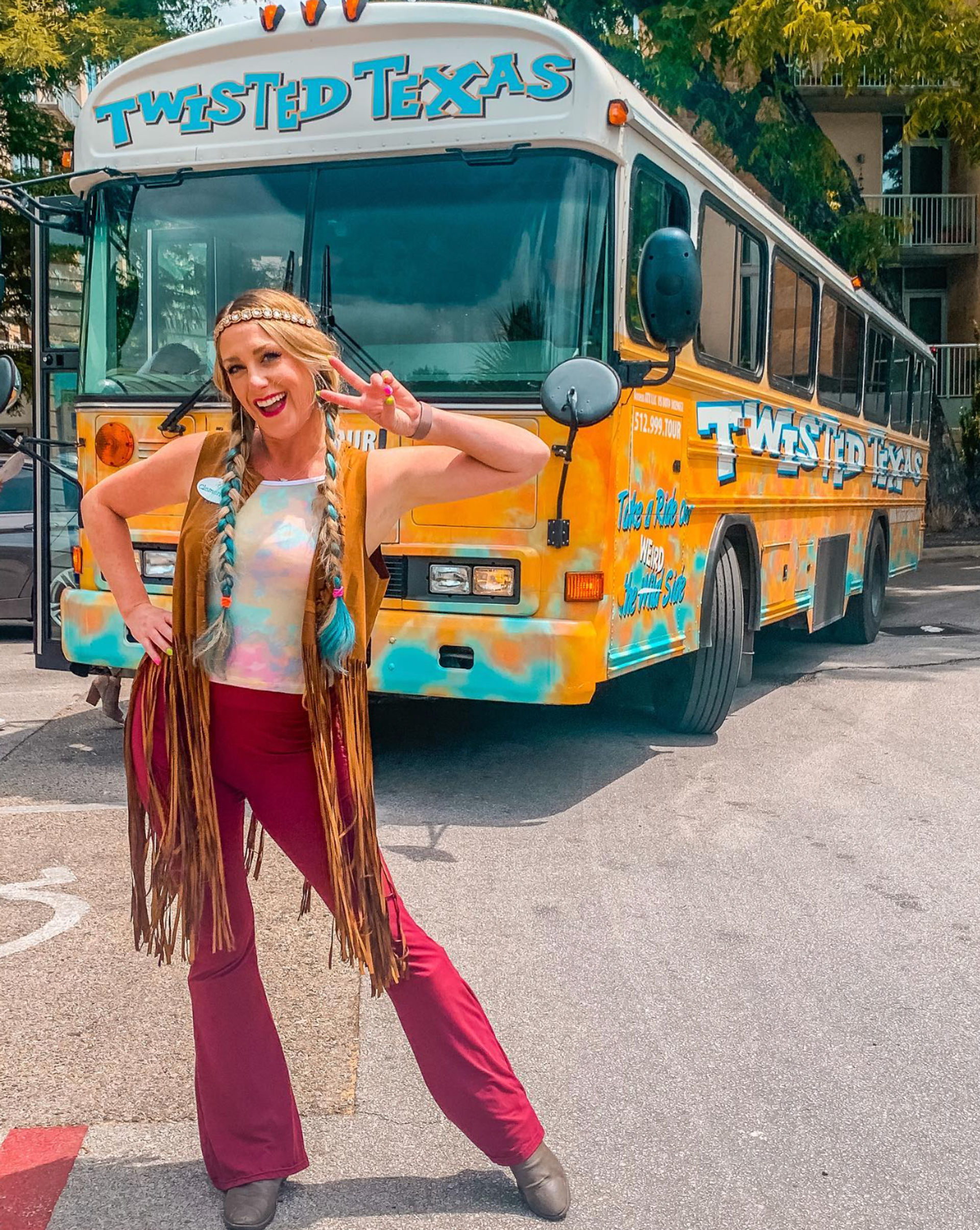 Woman in boho festival outfit with braided teal-tipped hair and fringe vest flashing a peace sign in front of a bright yellow-and-teal Texas tour bus parked on a sunny street