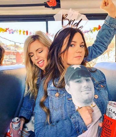 Two friends in denim jackets on a decorated party bus at a bachelorette celebration, one wearing a 'Bride' veil headband and holding a cardboard face cutout while both hold canned drinks.