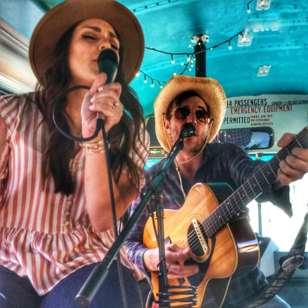 Acoustic duo performing live inside a decorated bus — female singer in a striped blouse and hat with a microphone, male guitarist in a straw hat and sunglasses playing an acoustic guitar under string lights.