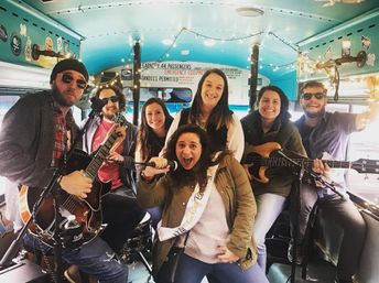 Lively group of seven friends playing guitars, bass and drums inside a turquoise party bus decorated with string lights, while a woman wearing a sash sings into a microphone for a bachelorette-style celebration.