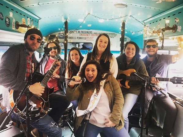 Lively group of seven friends playing guitars, bass and drums inside a turquoise party bus decorated with string lights, while a woman wearing a sash sings into a microphone for a bachelorette-style celebration.