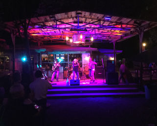 Outdoor wooden pavilion stage at night with a live rock band performing under purple and pink lights and a small audience watching