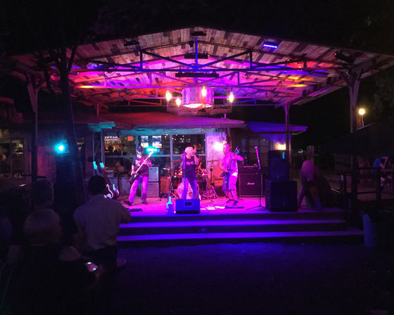 Outdoor wooden pavilion stage at night with a live rock band performing under purple and pink lights and a small audience watching