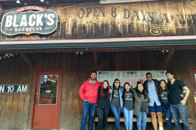 Group of adults smiling and posing in front of a rustic wooden barbecue restaurant storefront with a vintage sign and illuminated "open" signage