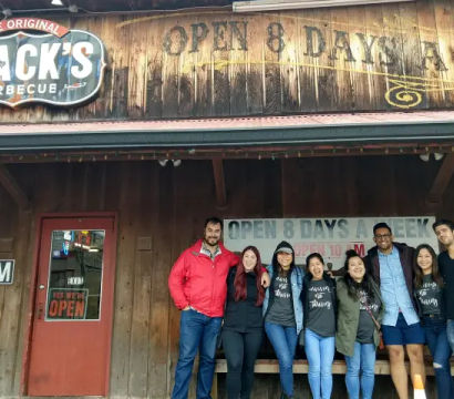Group of adults smiling and posing in front of a rustic wooden barbecue restaurant storefront with a vintage sign and illuminated "open" signage
