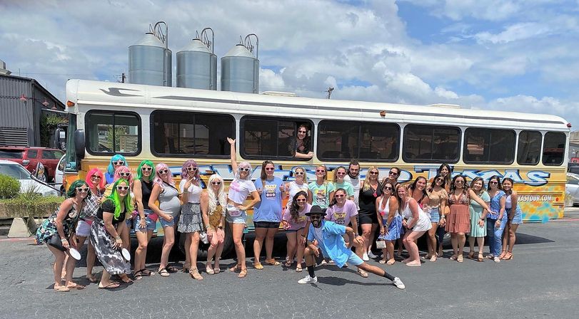 Large group of adults posing in front of a colorful tour bus in a sunny parking lot, many in sunglasses and bright summer outfits with industrial silos and blue sky in the background — fun road‑trip/tour vibe.