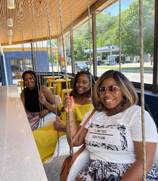 Three friends smiling on hanging swing seats at a bright window counter in a modern diner, sunny street and gas station visible outside.