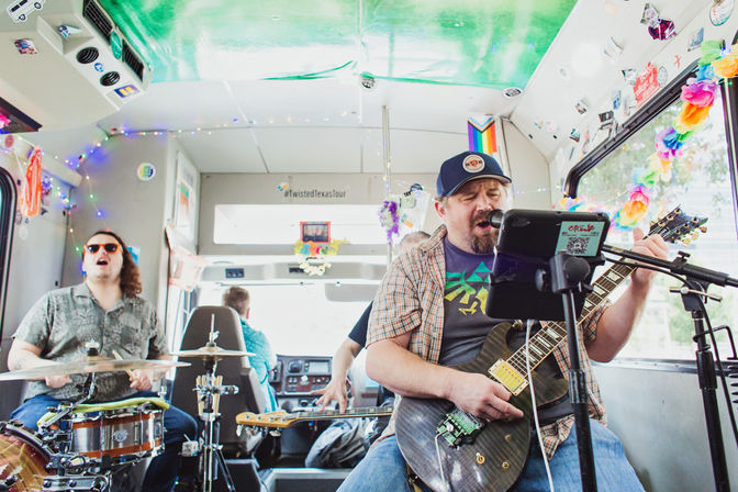 Indie band performing inside a decorated tour bus in Texas — singer with electric guitar at a microphone, drummer and bassist playing behind him, colorful string lights and rainbow leis lining the windows.