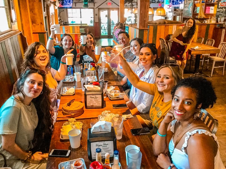 Smiling group of friends raising cups in a cheerful toast around a long wooden table during a casual brunch in a cozy rustic restaurant, plates, cups and condiments visible.